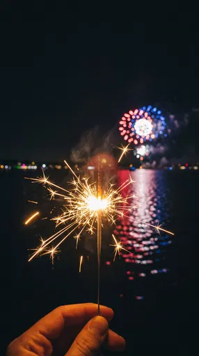 Hand Holding Lit Sparkler Near Waterfront Fireworks